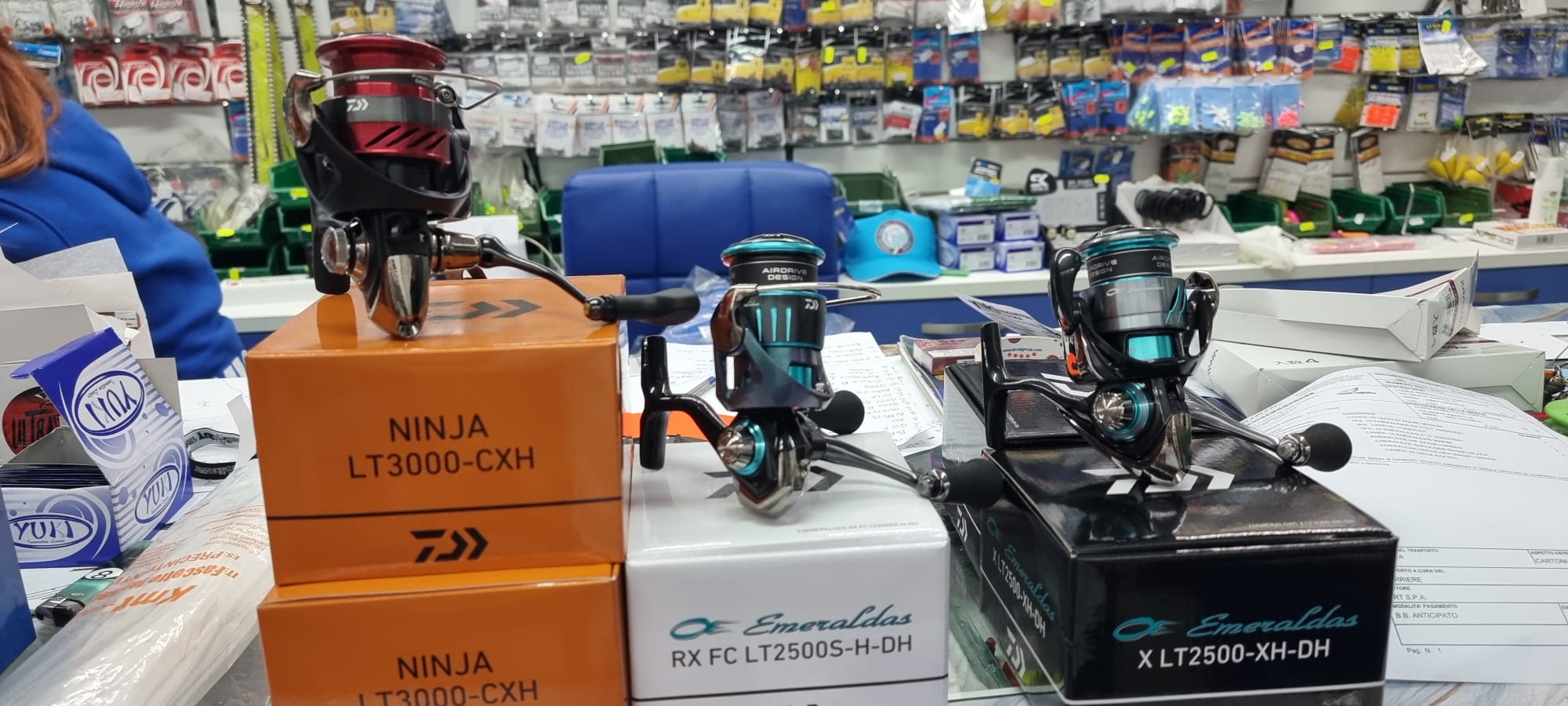 Mulinelli Daiwa Ninja rosso ed Emeraldas verde acqua sulle scatole, tra espositori di esche da L'Onda Perfetta