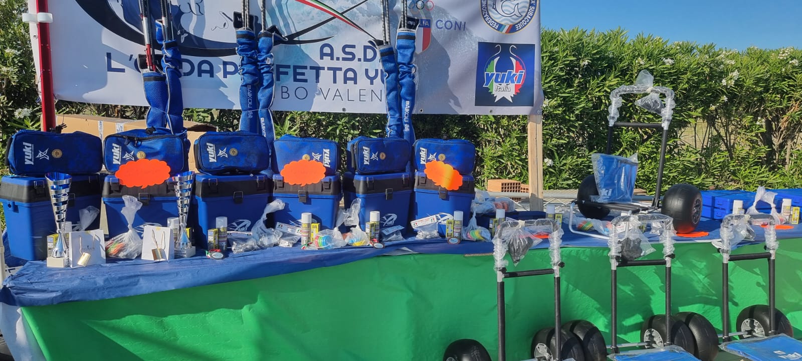Premi di gara di pesca: sedute portamateriale Yuki blu, borse portacanne, coppe e carrelli sotto lo striscione A.S.D. L'Onda Perfetta