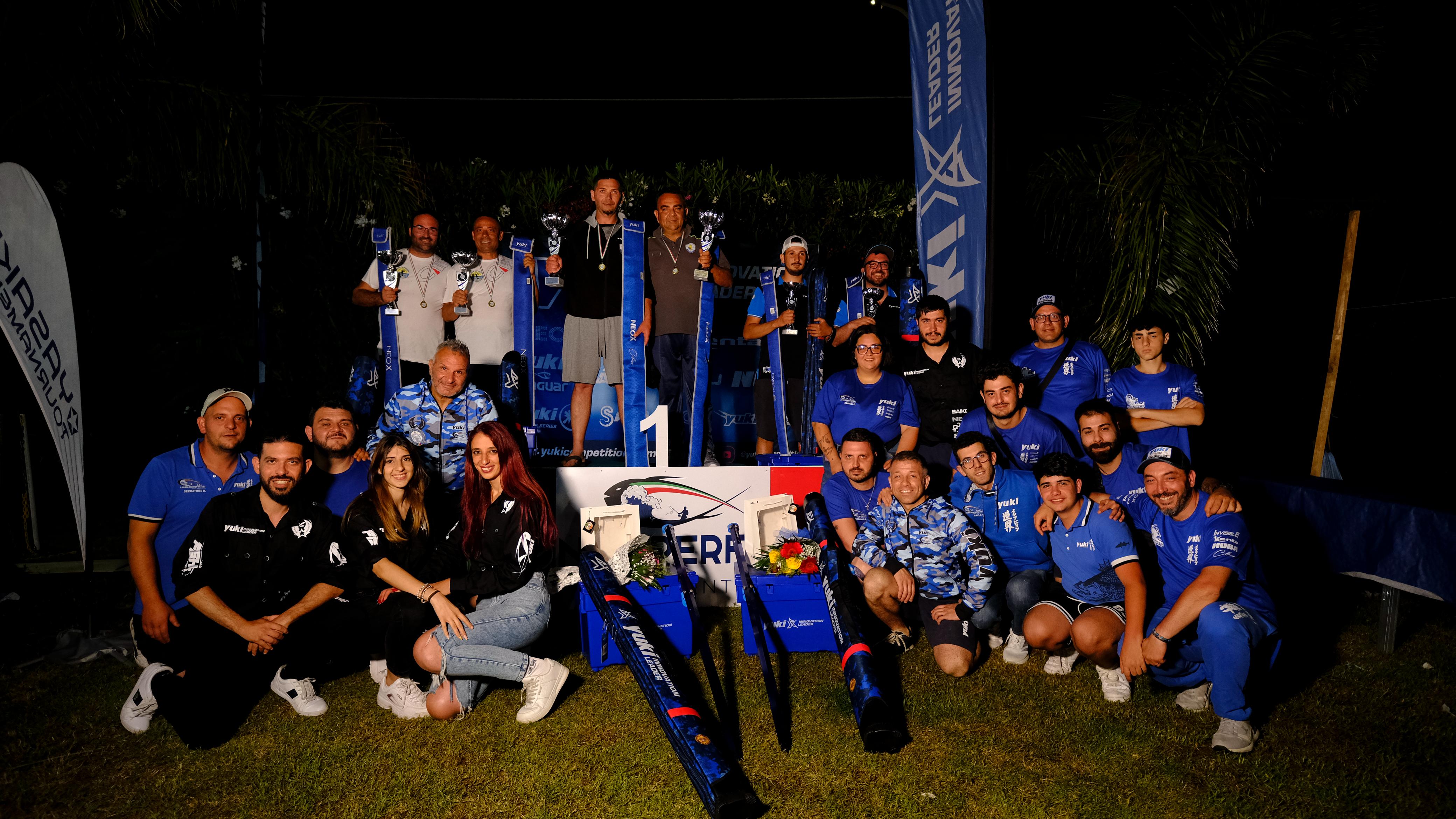 Pescatori in maglie blu Yuki posano di sera attorno al podio numero 1 con trofei, medaglie e foderi per canne
